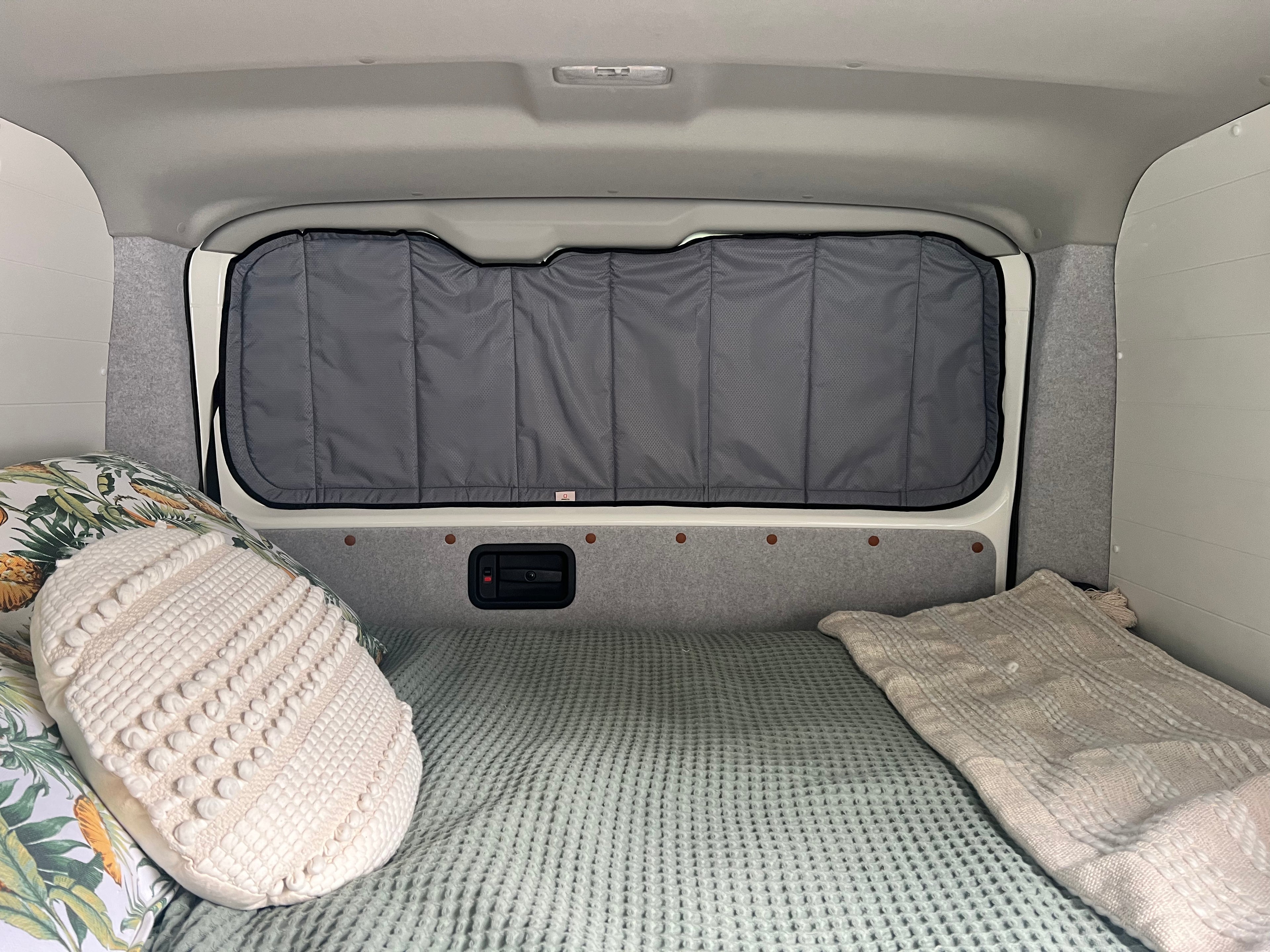 A dark gray padded cargo bed divider or privacy panel installed in the back of a vehicle, shown above a bed setup with a textured green blanket, cream pillow, and folded blanket.